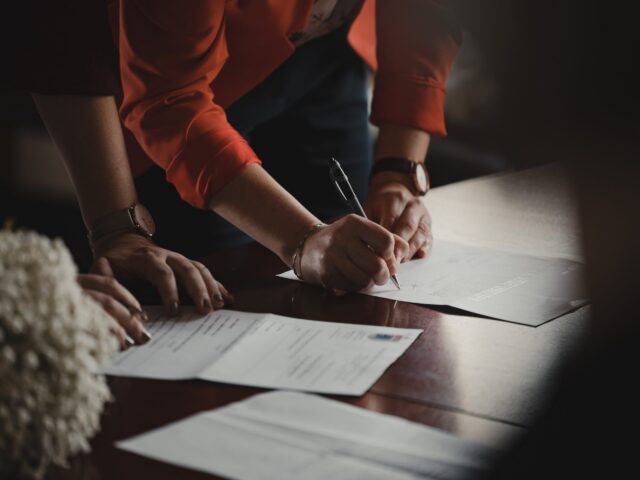 Two people are bent over a table, signing what appears to be a will. One person wears a red jacket and a watch on their left wrist. A bouquet of white flowers is partially visible in the foreground, adding an air of solemnity to the scene.