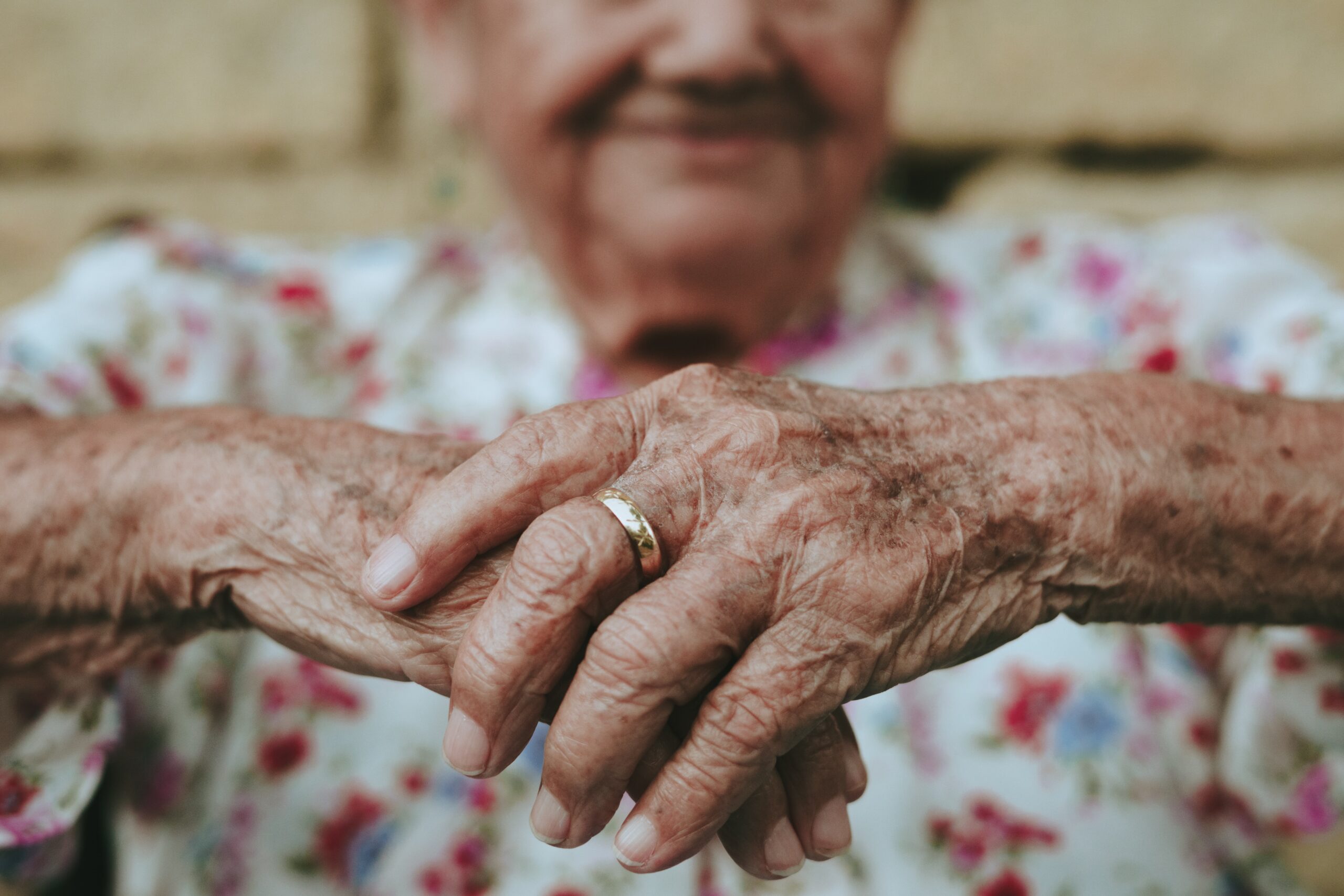 An elderly person with weathered hands, perhaps slightly ill, dons a ring on the left hand. Clad in a floral-patterned shirt, their face remains softly out of focus in the background.
