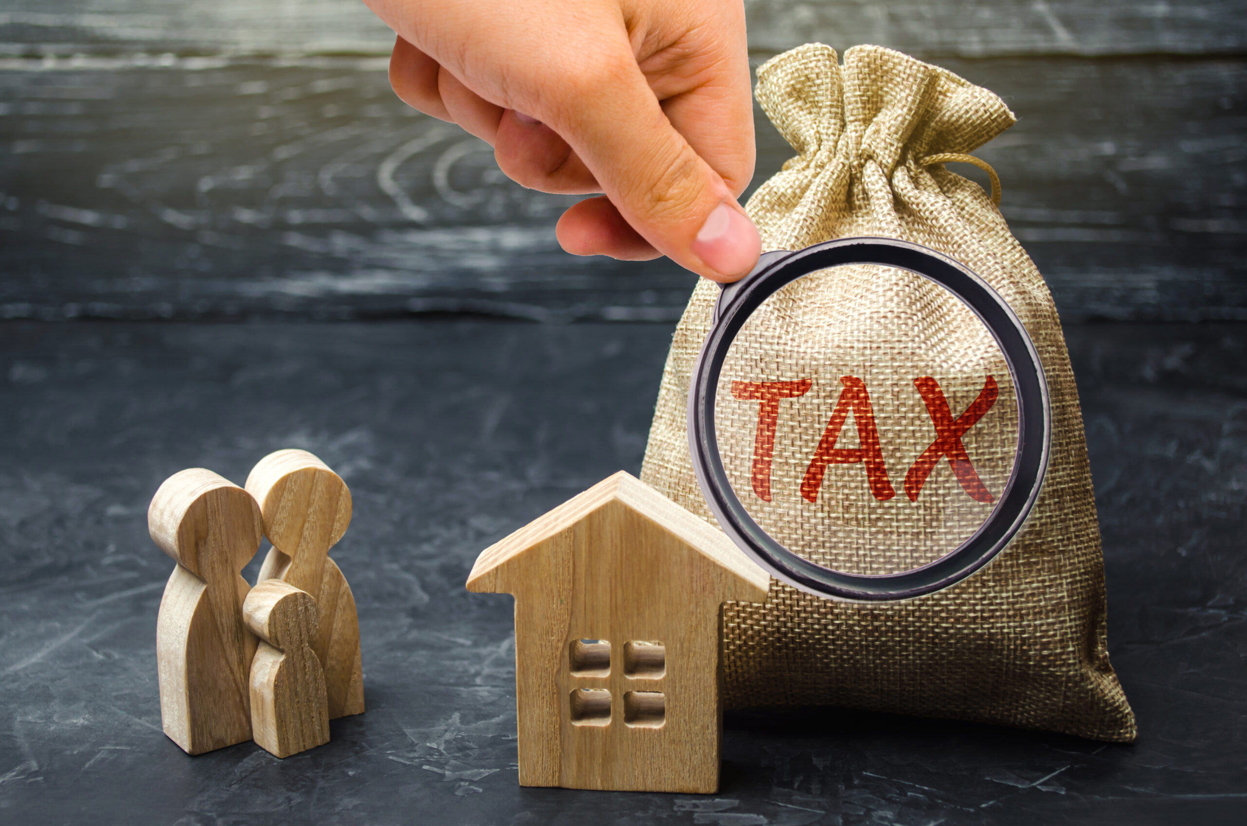 A hand holding a magnifying glass over a burlap sack labeled "TAX." In the foreground, there are wooden figurines representing a family next to a small wooden house model, all set against a dark background.