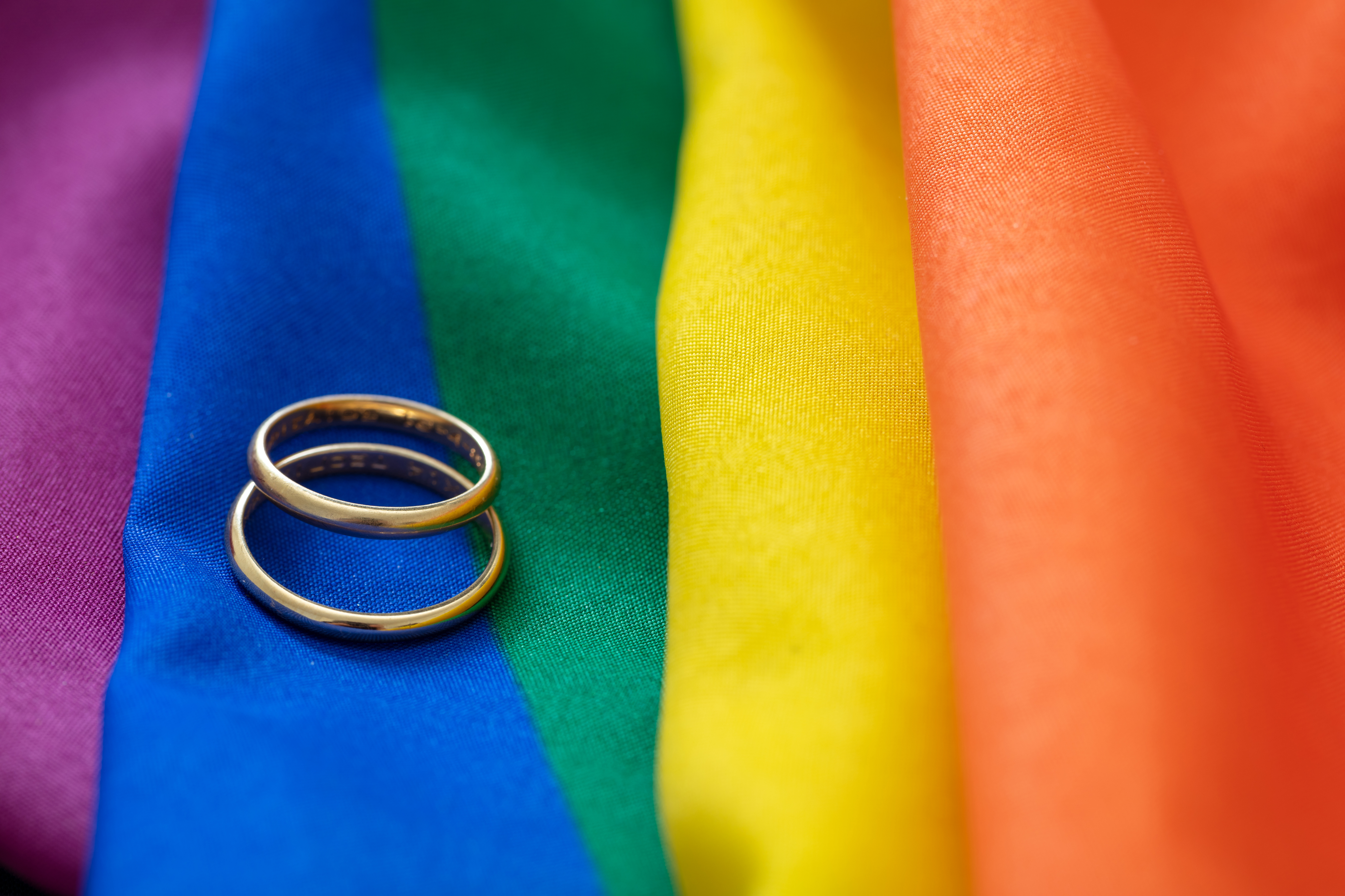 Two gold wedding rings rest on a rainbow-colored fabric, symbolizing LGBTQ+ pride and marriage equality.