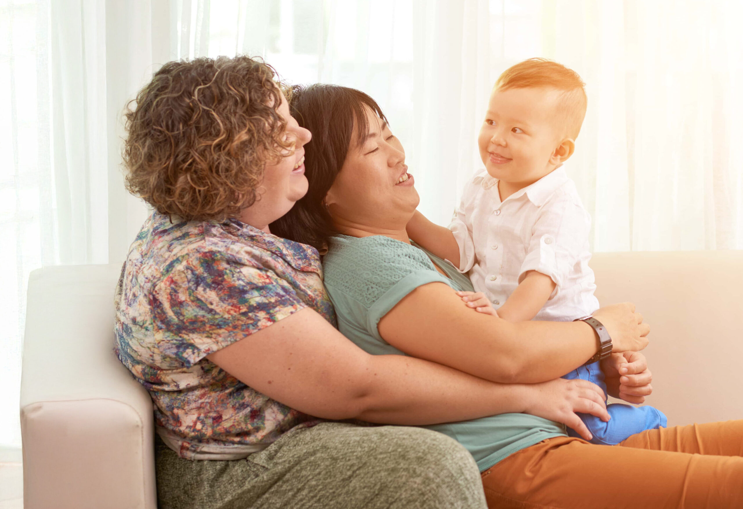 Two women sit close together on a couch, smiling and holding a young boy who sits on one woman's lap. The boy smiles warmly at the women as sunlight streams in. LGBTQIA+ Family Lawyers support families like theirs every step of the way.