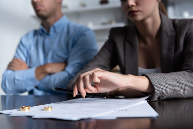 A man and woman sit at a table with divorce papers and two wedding rings in front of them; the woman holds a pen, while the man sits with arms crossed, both appearing serious as they navigate the divorce process.