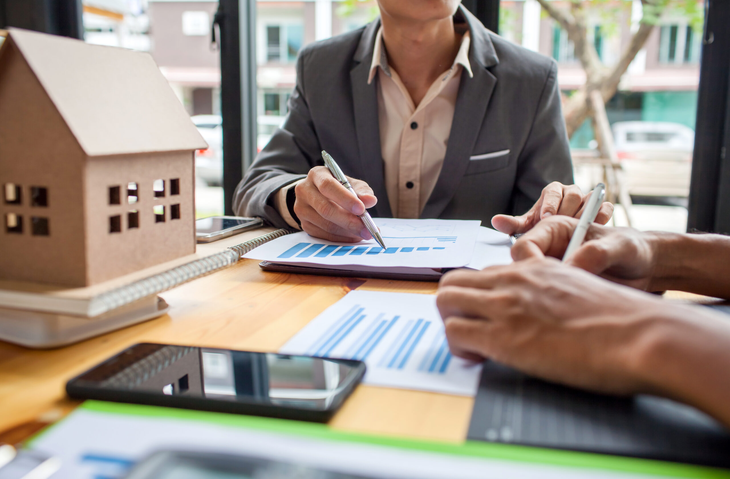 Two people in business attire are seated at a table with charts and documents. One holds a pen, pointing at a paper. A small model house is on the table, and a smartphone is nearby. A window with an outdoor view is in the background.