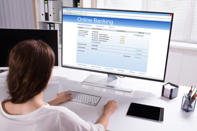 A person sitting at a desk uses a computer to check their online banking. The monitor displays account information, recent transactions, and a balance of $5,304.02. The desk has a tablet and paperwork next to the keyboard and mouse.