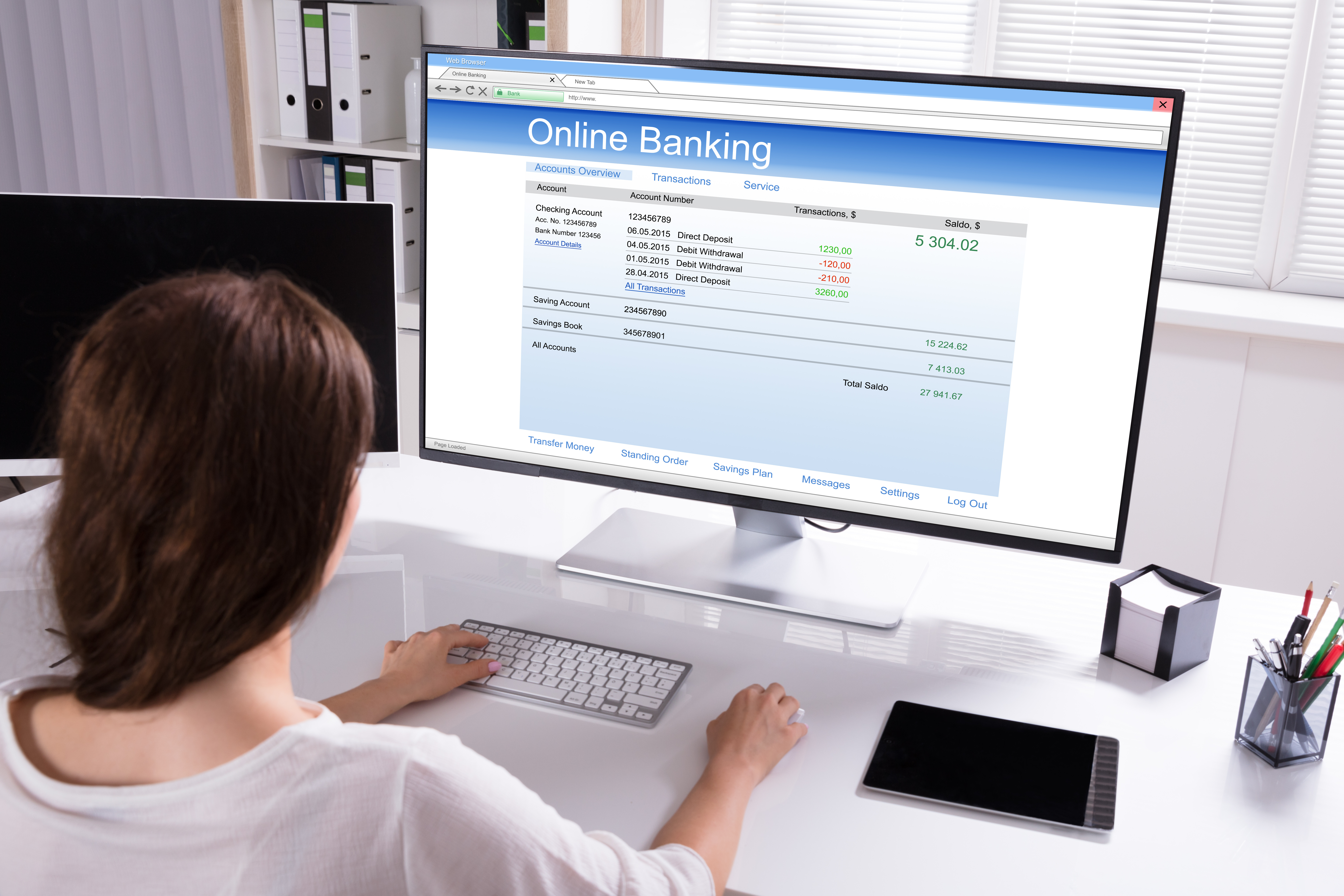 A person sitting at a desk uses a computer to check their online banking. The monitor displays account information, recent transactions, and a balance of $5,304.02. The desk has a tablet and paperwork next to the keyboard and mouse.