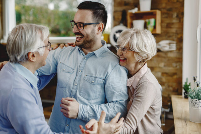 A joyful moment unfolds as three people, likely celebrating a decision to divide the estate fairly, smile and embrace indoors. An older man and woman stand on either side of a younger man in a light blue shirt. The room exudes a warm, cozy atmosphere with wooden accents and plants.