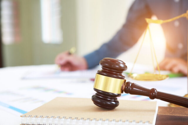 A gavel rests on a notebook in the foreground, while in the background, a person meticulously writes on a document—a transfer-on-death deed—beside scales of justice, all elegantly arranged on a white table bathed in natural light.