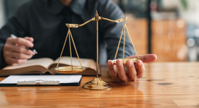 A person sits at a wooden desk with an open book and clipboard, holding a golden balance scale symbolizing justice or law. The background is blurred.