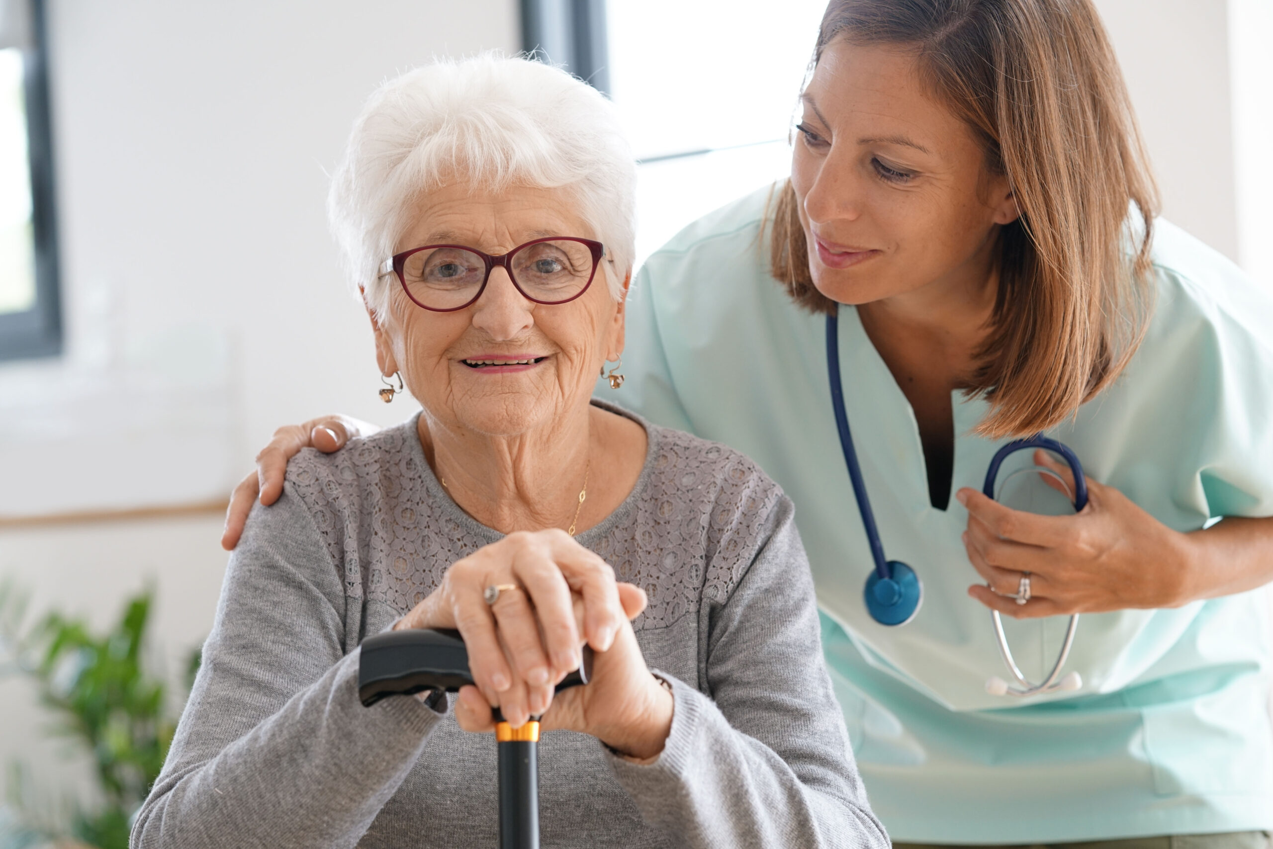 An elderly woman with white hair and glasses smiles while holding a cane, as a caring healthcare worker in scrubs and a stethoscope gently places a hand on her shoulder—reflecting trust that goes beyond nursing home costs.