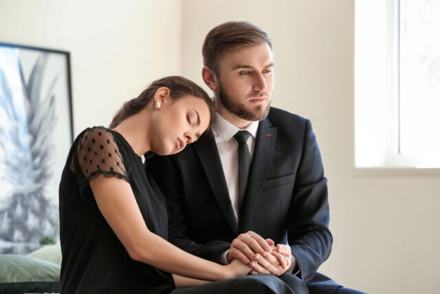 A woman in a black dress rests her head on a man’s shoulder. The man, dressed in a black suit and tie, holds her hand. Both have somber expressions, sitting together in a brightly lit room.