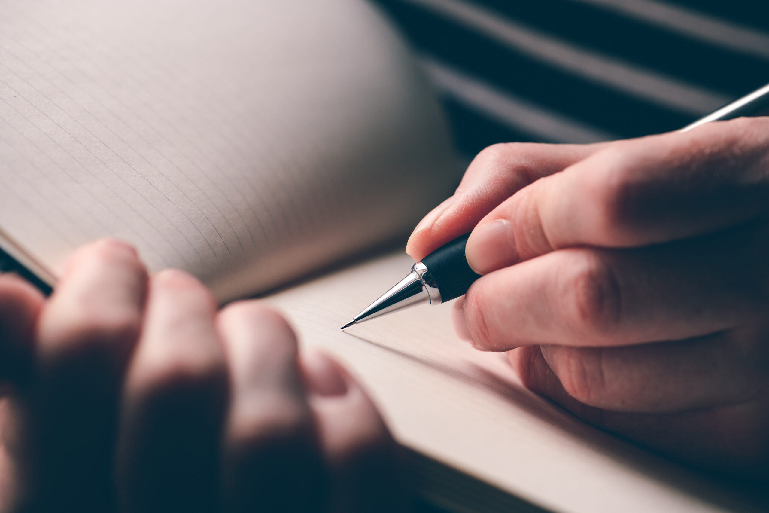 A close-up of a hand holding a mechanical pencil, poised to write a handwritten Will on the blank page of an open notebook with faint grid lines. The background shows the blurred outline of the writer’s other hand.