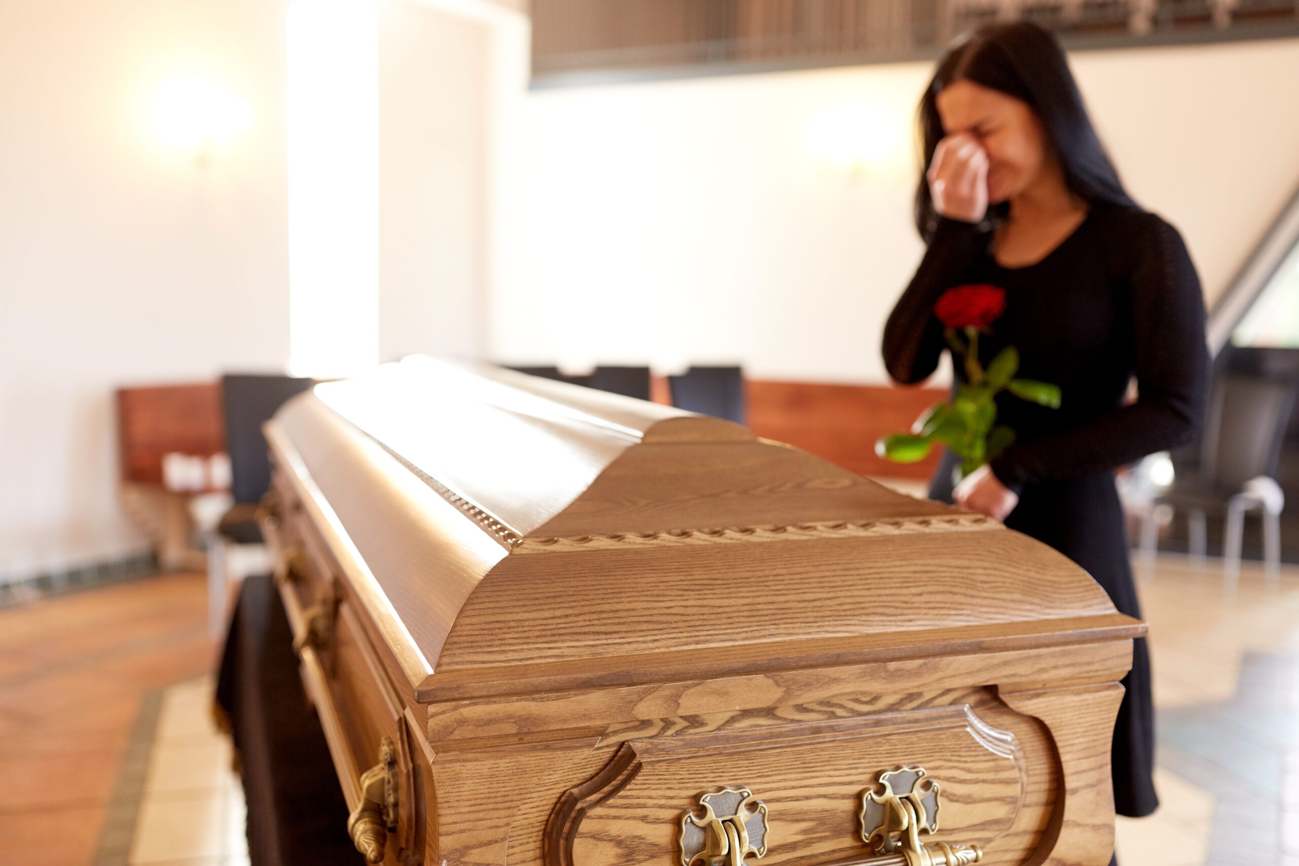 A woman in black, holding a red rose, stands by a wooden casket and wipes tears from her face in a bright room, indicating a funeral or memorial service.