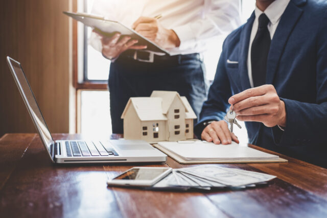 Two people in business attire sit at a desk with a laptop, clipboard, model house, cash, and keys. One holds house keys while the other writes on a clipboard, indicating a real estate trust transaction.