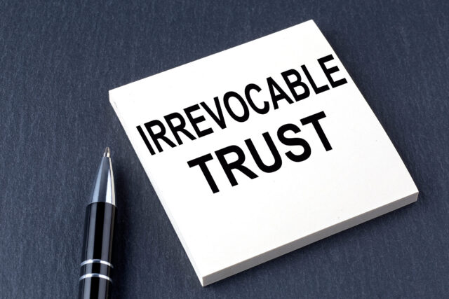 A black pen rests beside a white notepad labeled "IRREVOCABLE TRUST" in bold black letters, highlighting the importance of an irrevocable trust against a sleek, dark background.