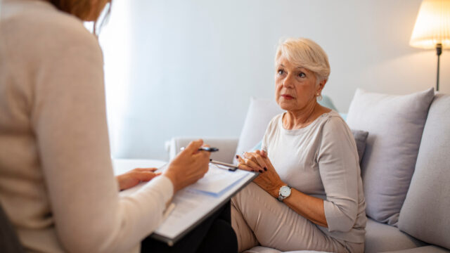 An older woman sits on a couch, attentively listening to a person with a clipboard and pen, discussing concerns about a will contested for undue influence in a calm, well-lit room.