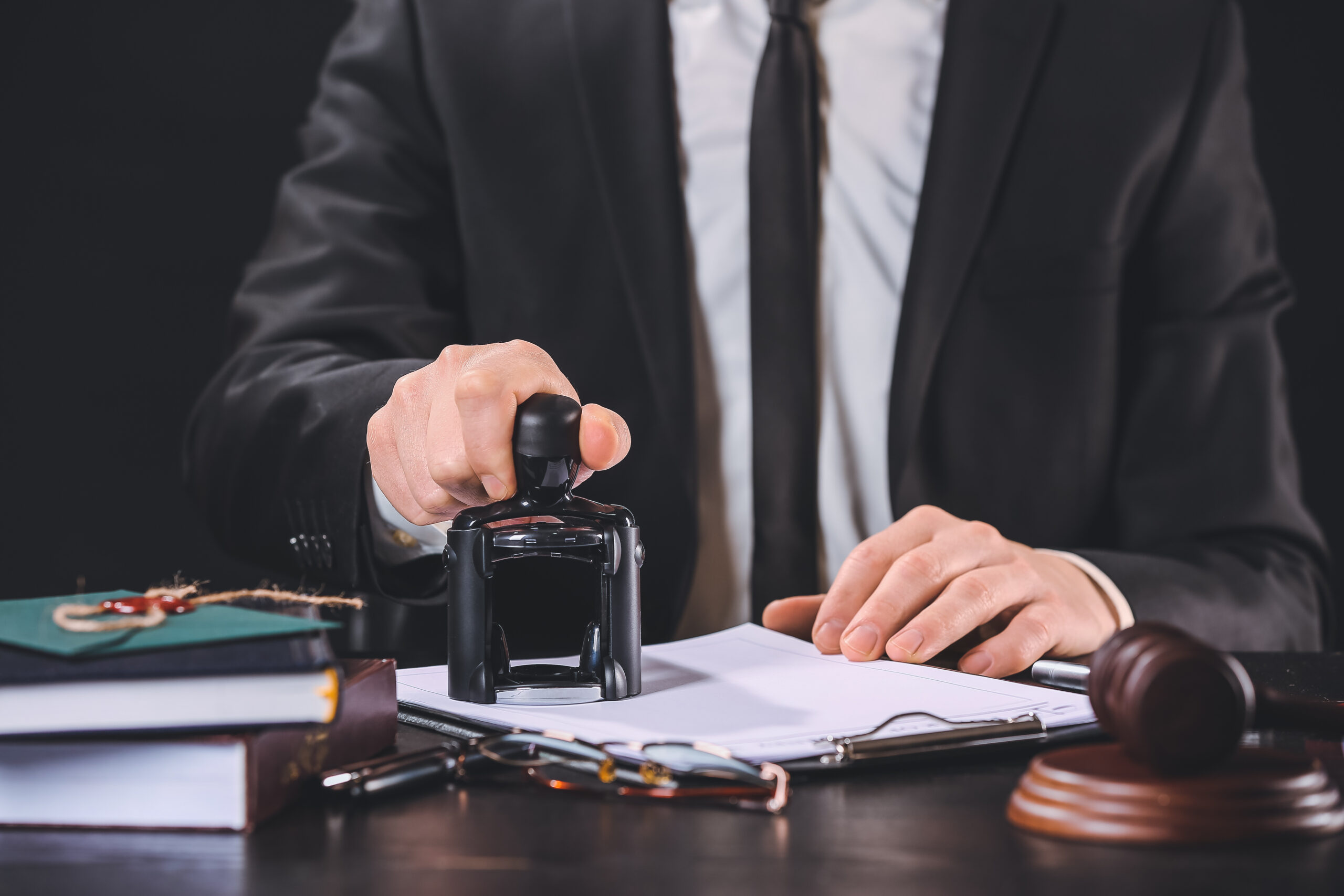 A person in a suit stamps a document on a desk with books, glasses, a pen, and a judge's gavel, suggesting a legal or official setting.