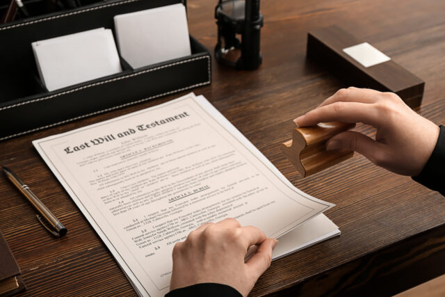 A person stamps a last will and testament document on a wooden desk, surrounded by a pen, letter organizer, and other office supplies.