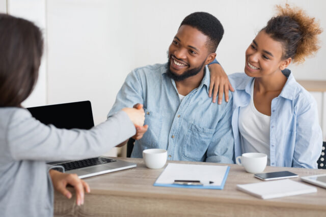 A smiling couple sits at a desk, shaking hands with a professional across from them. Papers, a clipboard, coffee cups, and a laptop are on the table, hinting at a business meeting discussing how wills can override beneficiary designations.