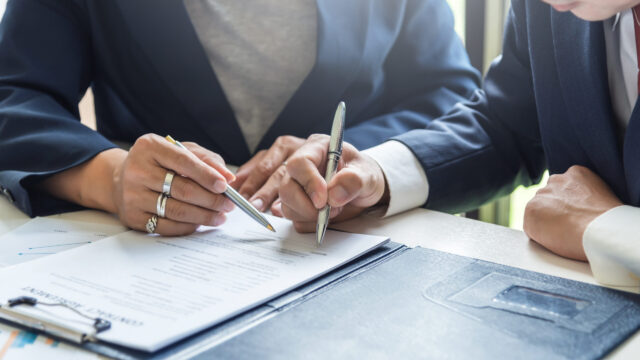 Two people in business suits sitting at a desk, reviewing and discussing a document. One person points at the paper with a pen while the other prepares to write or sign. A folder and paperwork are visible on the desk.