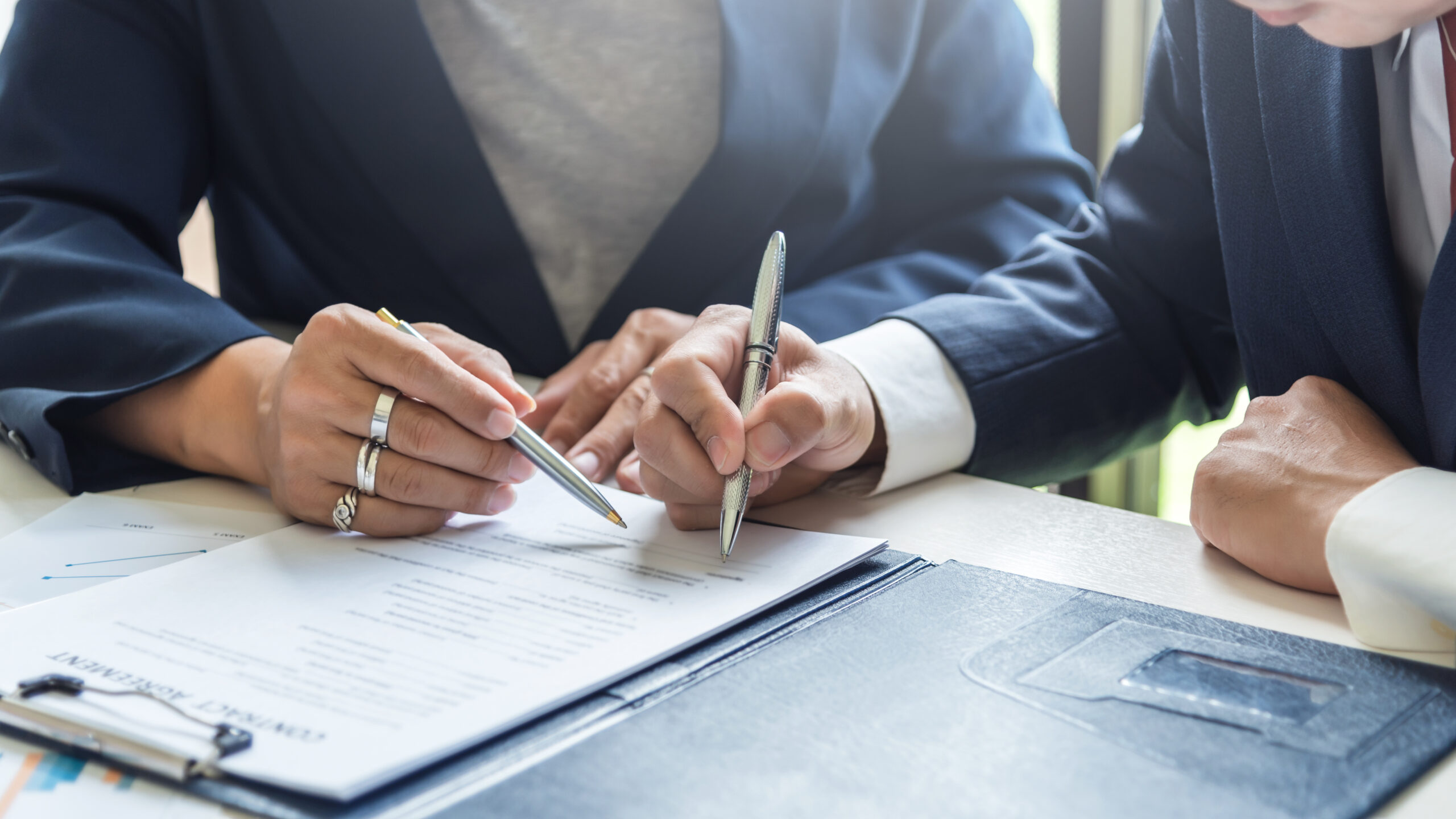 Two people in business suits sitting at a desk, reviewing and discussing a document. One person points at the paper with a pen while the other prepares to write or sign. A folder and paperwork are visible on the desk.