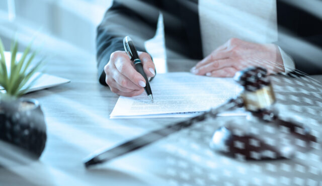 A person in business attire signs a document at a desk, with a gavel and small plant nearby, suggesting a legal or professional setting where establishing guardianship may be necessary.