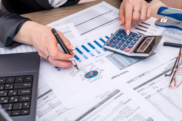 A person using a calculator and holding a pen is working with financial documents and charts on a desk, alongside a laptop, stapler, and eyeglasses.
