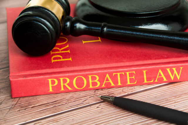A black judge’s gavel rests on top of a red book titled "PROBATE LAW" in gold letters, with a black pen lying nearby on a wooden surface.