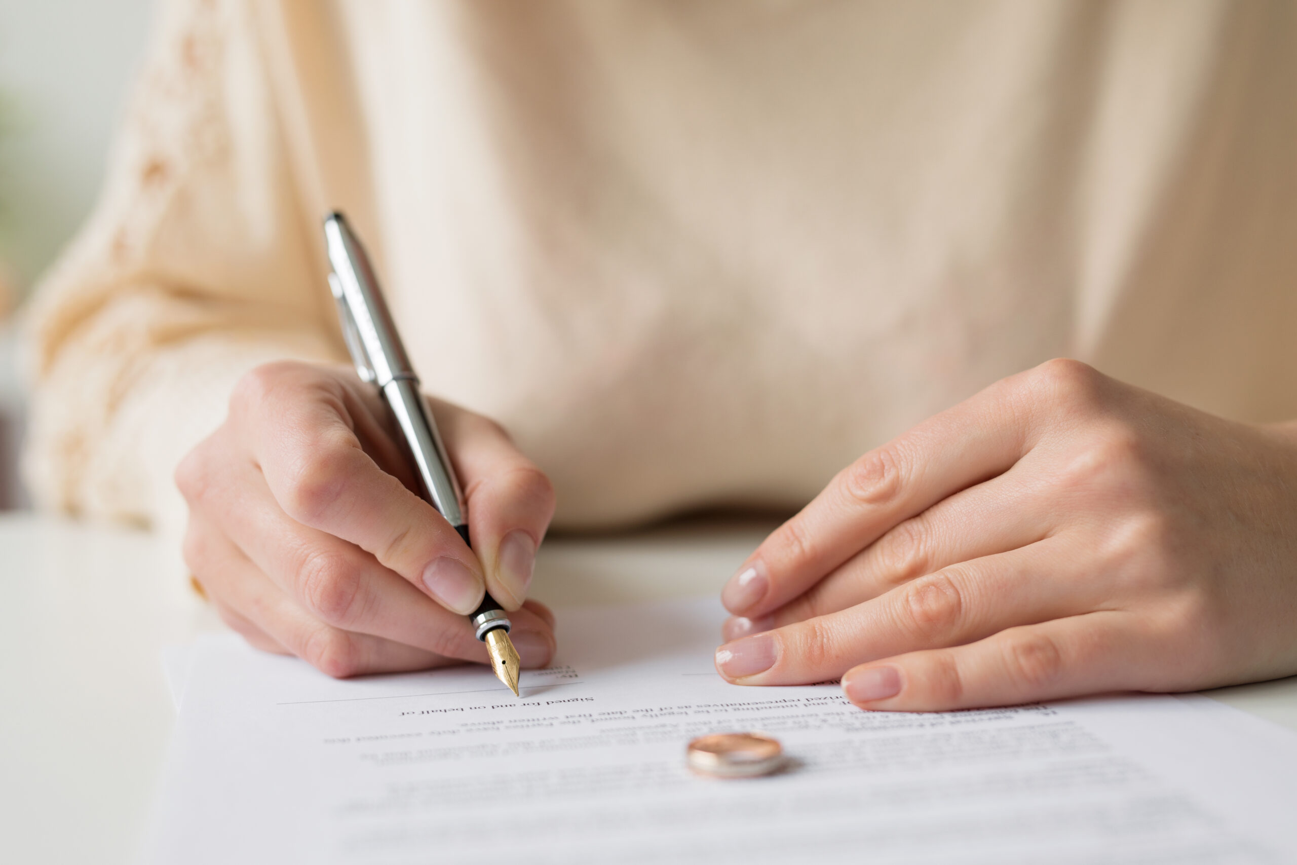 Close-up of a person signing a document with a pen, a wedding ring placed on the paper—emphasizing how will beneficiary designations can be affected by divorce. The focus is on their hands and the document.