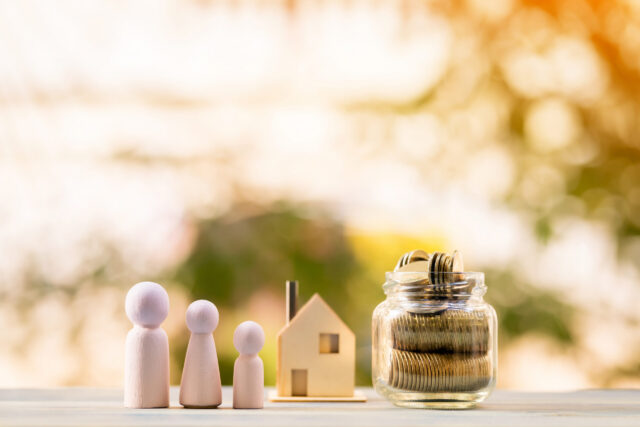 Wooden figures of three people, a small house model, and a glass jar filled with coins are arranged on a table outdoors, symbolizing family, home, and saving money. The background is blurred with warm sunlight.