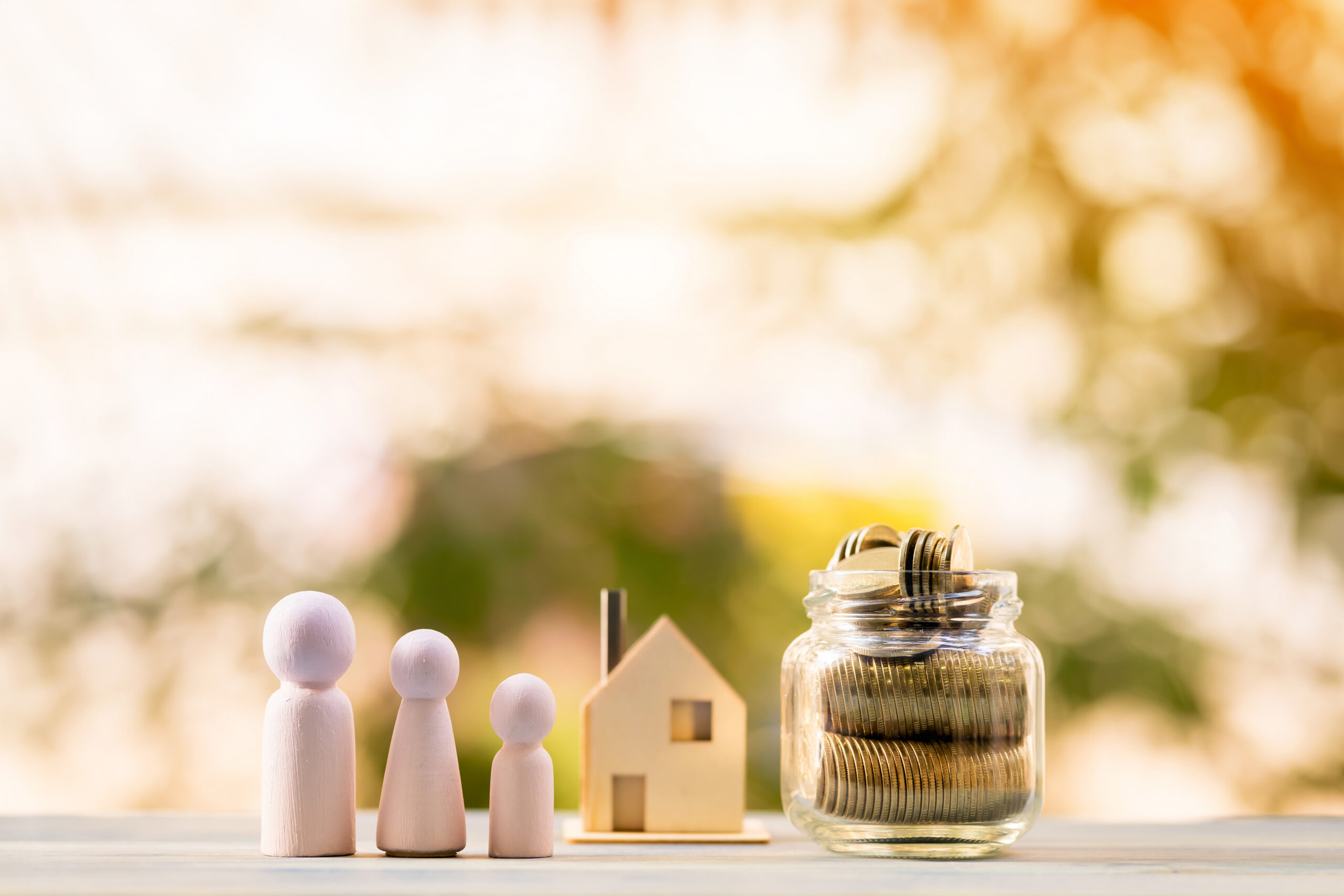 Wooden figures of three people, a small house model, and a glass jar filled with coins are arranged on a table outdoors, symbolizing family, home, and saving money. The background is blurred with warm sunlight.