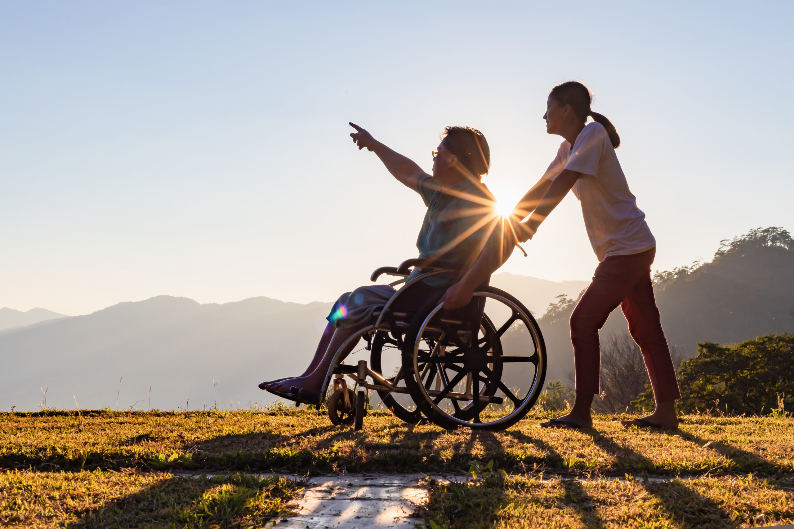 A person in a wheelchair points toward the sky while another stands behind, pushing them on a grassy hill at sunset with mountains in the background—an inspiring scene that highlights the importance of disabled estate planning.