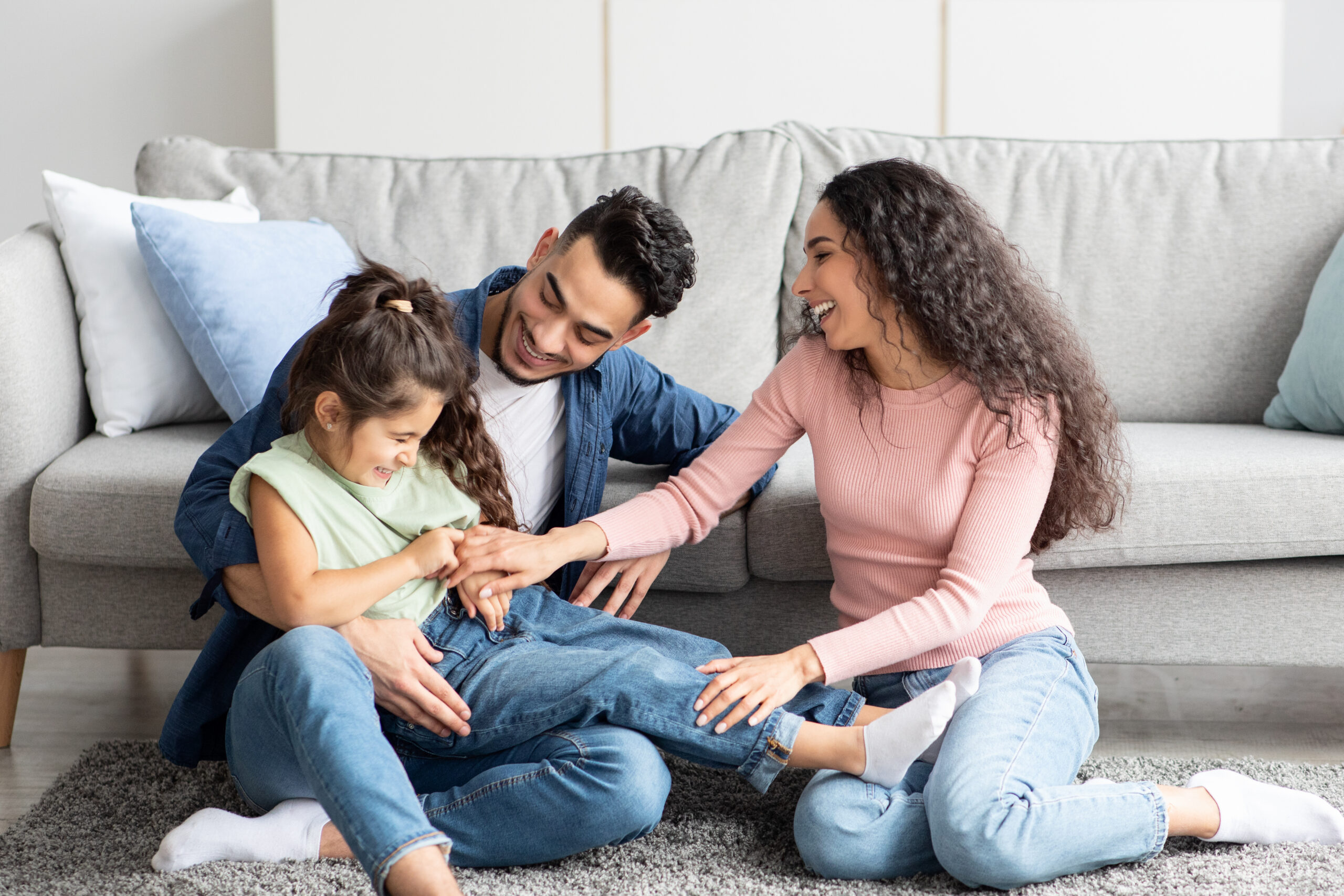 A cheerful family of three sits on a carpet in a living room, with the parents tickling and playing with their young daughter, all smiling and laughing together. A gray sofa with cushions is in the background.